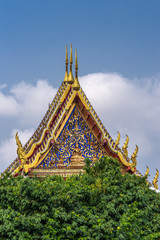 Bangkok city, Thailand - March 17, 2019: Three golden gables with intense blue decoration towering over green foliage at Temple of Dawn against blue sky.