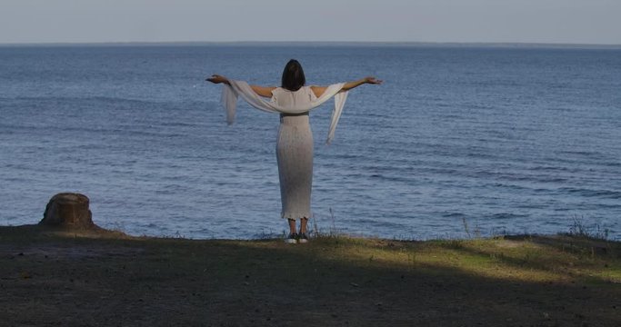 Back view of a charming Caucasian woman in light white dress standing on the background of waves with stratched out hands. Cheerful girl spending summer day on the beach.