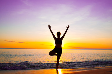 A young lady practices yoga on the beach in front of the setting sun. The Caribbean sea is tranquil as it laps the sandy coastline of the idyllic island