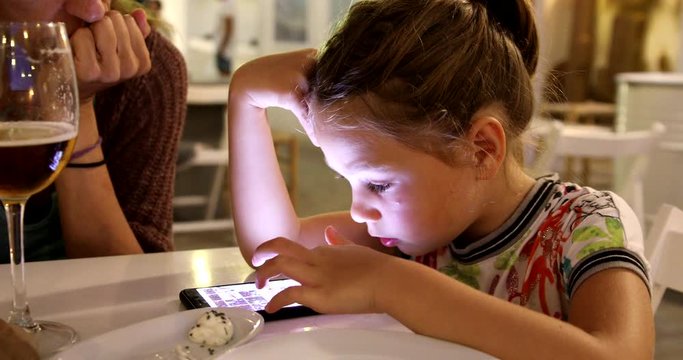 Portrait of six years old cute child with white dress, using smartphone, dinning in a restaurant next to her mother
