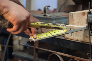Working in a workshop a blue collar laborer, solo man with tough rough skin on hands uses a folded metal measuring tape to find the dimensions of a piece of steel before cutting