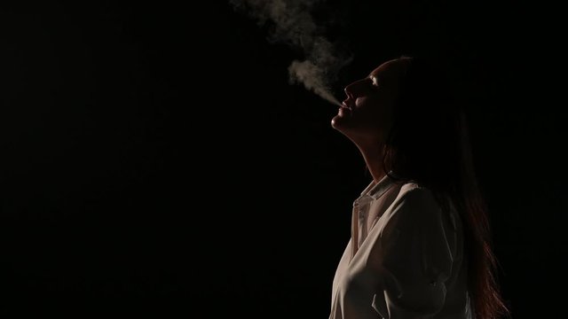 Pensive brunette in a white shirt smokes a hookah in the dark. Portrait of a woman smoking a vape, hovering. Black background. Electronic Cigarette. Bad habit.