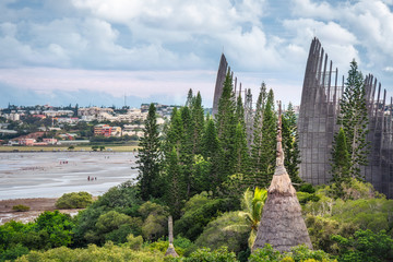 Tjibaou Cultural Centre, the Kanak native art museum with a traditional straw hut in foreground and the city of Noumea in background and people walking on sea shore at low tide in New Caledonia.