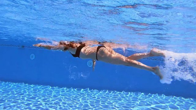 underwater side view following redhead woman with black bikini swimming crawl style in clear water of pool 