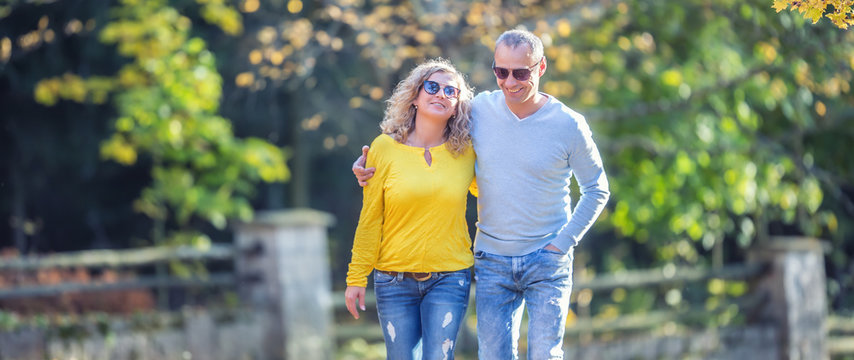 Happy Mature Couple In Autumn Park In The Embrace Walk On The Fallen Maple Leaves