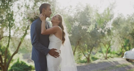 Smiling diverse couple embracing and kissing in nature on their wedding day with a vintage car in the background, happy multi ethnic bride and groom sharing a moment together