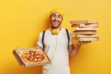 Delivery concept. Man pizza dealer holds stack on cardboard boxes, shows tasty fast food in opened container, works as courier, wears yellow hat and white t shirt, uses headphones for listening audio.