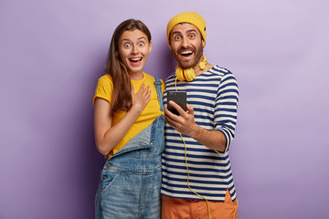 Naklejka premium Joyful female and male teenagers hold smartphone gadget, dressed in stylish apparel, type message, watch video content in social networks, check notification, have happy mood, enjoy leisure time