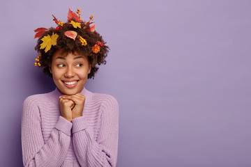 Tender feminine woman keeps hands under chin, spends good time with friend, expresses positive emotions after autumn walk, has foliage in curly hair, chuckles from hilarious joke, dressed in sweater