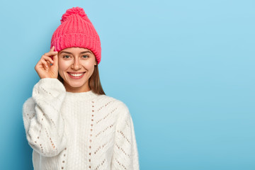 Good looking brunette young woman keeps hand near face, smiles pleasantly, wears hat and white knitted sweater, enjoys pleasant moment, expresses good emotions, poses against blue studio wall