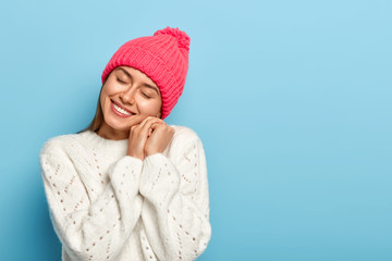 Cheerful European woman recalls nice pleasant memories, tilts head, keeps hands near face, smiles happily, wears rosy knitted hat and white sweater, poses over blue background. Tenderness and beauty