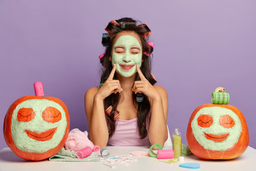 Portrait of pleased young woman keeps both fore fingers on face, shows natural clay mask, wears...