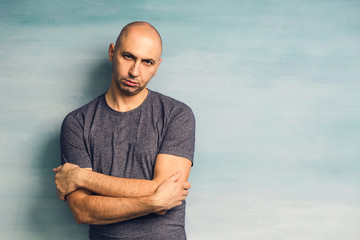 A young bald man in a gray T-shirt on a blue background looking angrily at the camera,
