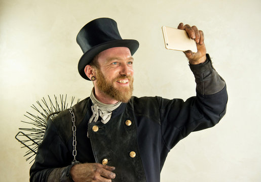 Schornsteinfeger mit Zylinder, in Arbeitskleidung und Kehrbesen, macht ein Selfie mit Mobiltelefon