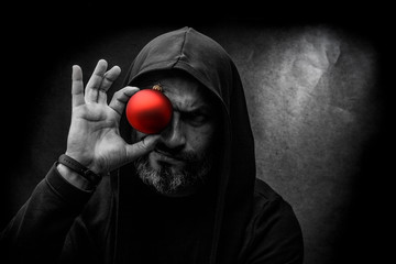 Black and white portrait of a bald bearded man in a hood with red christmas ball on a dirty gray background. Young Santa concept.