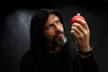 Portrait of a bald man with a beard in a black hood witha red christmas ball on a dirty gray background. Young Santa concept.