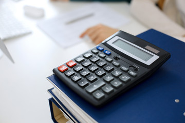 Calculator and binders with papers are waiting to be processed by business woman or bookkeeper working at the desk in office back in blur. Internal Audit and tax concept