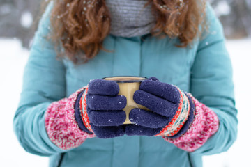 Woman’s hands in knitting colorful gloves are holding metal cup with hot tea in winter park.