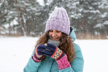 Beautiful smiling woman with long curly hair in blue winter jacket and knitted colorful gloves is holding in her hands cup of hot tea on a walk in winter snowy forest.