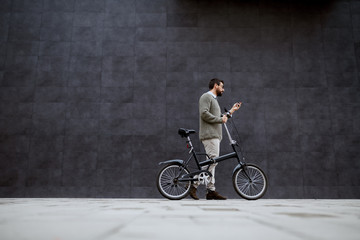 Fototapeta na wymiar Full length of stylish caucasian man pushing bicycle and using smart phone while passing by gray wall.