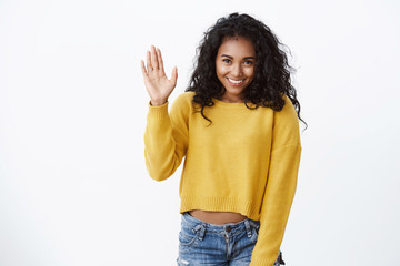 Shy lovely african-american girl in yellow sweater, curly hairstyle, stooping as raising hand,...