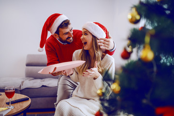 Fototapeta premium Amazed cute Caucasian blonde woman sitting on sofa in living room and receiving gift from her boyfriend. Both having santa hats on heads. In foreground is christmas tree. Living room interior.