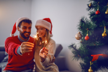Happy handsome caucasian couple with santa hats on heads sitting on sofa in living room,celebrating...