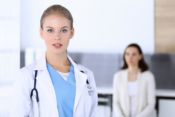 Doctor woman filling up medication history record while standing in emergency hospital office with patient in queue at background. Physician at work, portrait shoot. Medicine and health care concept