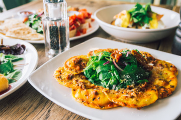 Chef dish of porcini mushroom ravioli on a wooden table with eggs benedict and plater of wings with bruschetta in the background.