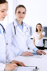 Fototapeta premium Group of doctors at work in hospital with patient in a queue at the background. Physician filling up medical documents or prescription while standing at reception desk. Data in medicine and health
