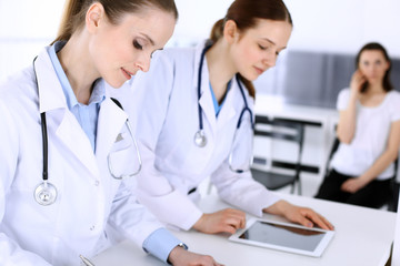 Fototapeta premium Group of doctors at work in hospital with patient in a queue at the background. Physician filling up medical documents or prescription while standing at reception desk. Data in medicine and health