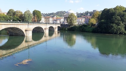 Fototapeta na wymiar Torino, Italy - 10/17/2019: Beautiful view to the river Po, with an amazing reflection of the houses and the bridge on the water. Clear blue sky and autumn coloured trees in the background. 