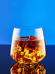 Crystal glass with a double portion of whiskey on ice on a glass surface and a blue background. A glass of whiskey is reflected on the surface.
