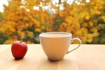 cup of cocoa with milk or cappuccino and a red apple on a table with a view of yellow and orange trees, autumn mood concept, close-up