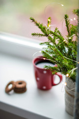 Cozy time. Fir branches in a vase on the window, lights garlands. Red Cup of coffee and cookies in focus.