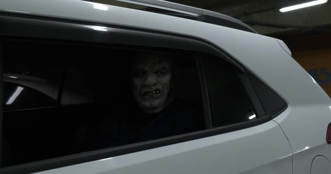 In back seat of white car sits maniac in white scary mask, wry smile, scars, scares passers-by, Halloween, terrible costume of killer from psychiatric hospital, dark parking lot, horror from the dark