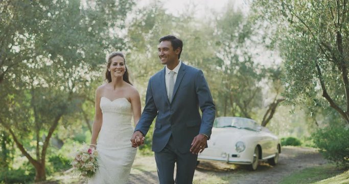 Happy smiling diverse couple running together holding hands on their wedding day with green trees and a vintage car in the background, multi racial bride and groom eloping together
