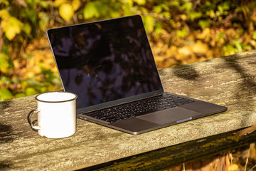 A laptop with an open lid stands on a wooden table in the autumn garden. Nearby is a white enameled metal mug. The table top is covered with small lichen. Yellow fallen foliage in the background. - Powered by Adobe