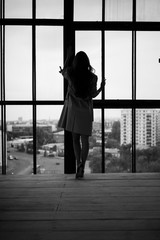 Silhouette of a woman in a trench coat in heels, the female looks at the city through the glass of a high-rise building, lonely and sad locked up