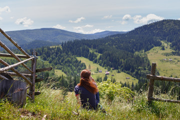 Fototapeta na wymiar Young redhead woman sitting on the hill and thinking, looking at summer mountains nature, resting after climbing and hiking - healthy lifestyle, travel, weekend trip, green tourism concept