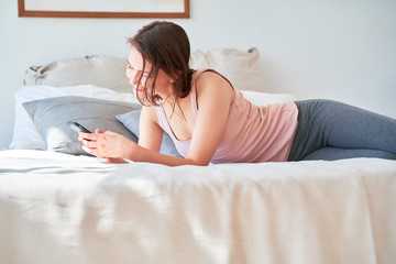 Image of young brunette with phone in her hands lying on bed