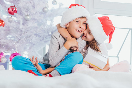 Two kids wearing santa hats and pyjamas and sharing christmas gift boxes in christmas designed atmosphere.