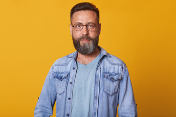 Obraz premium Photo of attractive middle aged man with serious and calm facial expression, having beard, dresses casual denim jacket and eyewear, posing isolated over yelow studio wall. People emotions concept.