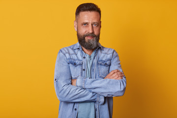 Obraz premium Representative, personable man with beard looking directly at camera, stands with arms folded, isolated over yellow background, dresses stylish denim jacket, having seriousfacial expression.