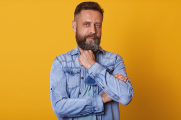 Fototapeta premium Serious thoughtful clever middle aged male wearing denim jacket, having beard, tries to remember something. Pensive unshaven man touching her beard with hand, isolated over yellow studio wall.