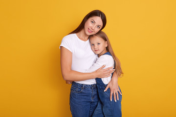 Close up portrait of beautiful young mother and her charming little daughter, stand smiling against yellow studio wall, woman hugging her child with great love, wearing white shirt and denim trounsers