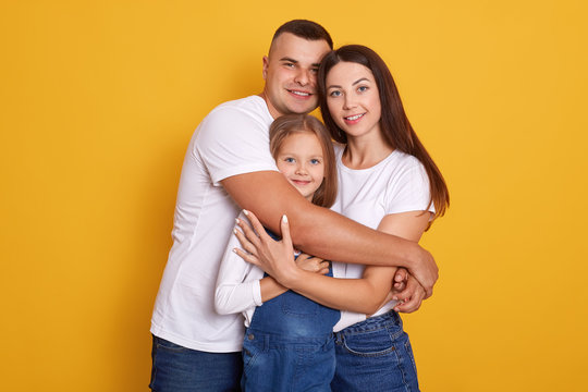 Horizontal Shot Of Beautiful Young Family Hugging, Looking Directly At Camera And Smiling While Standing Against Yellow Studio Wall, People Dress Casually, Expressing Happyness. Relationship Concept