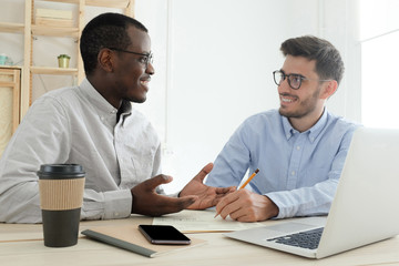 Teamwork concept. Diverse smiling modern businessmen using laptop together, caucasian and african coworkers at white office