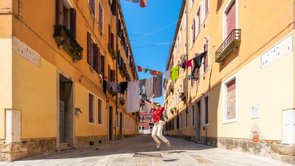 Fototapeta na wymiar Girl traveler in Venice. A joyful asian woman in red is jumping on Venice street. Colorful laundry is dried on the clotheslines between the houses. Authentic Venetian street in sunshine. Tourism Italy