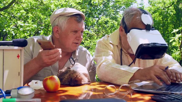 aged man with smartphone holds hedgehog and talks to senior citizen in headset typing on laptop. Concept virtual reality